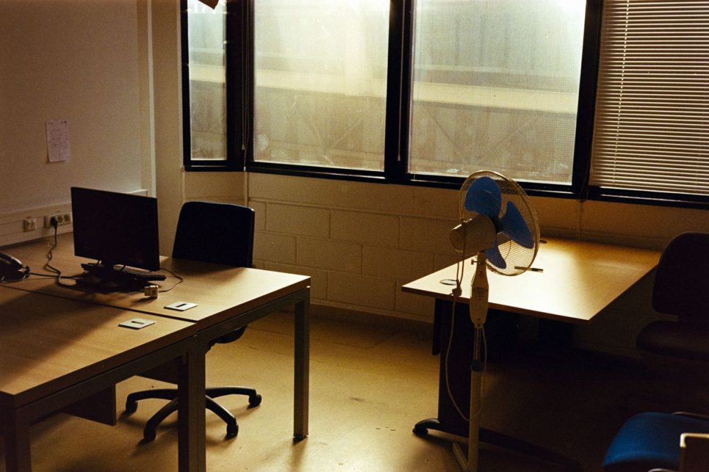 Quiet empty office with a desk, computer, and fan under warm afternoon sunlight.