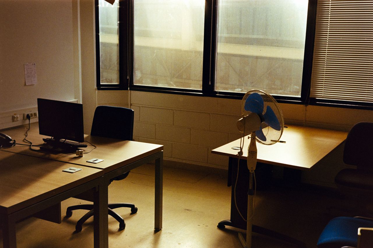 Quiet empty office with a desk, computer, and fan under warm afternoon sunlight.